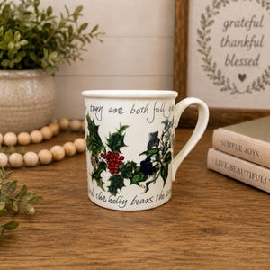 Könnte beinhalten: Weißer Keramikbecher mit Henkel, verziert mit Stechpalmenblättern, roten Beeren und Text. Der Becher trägt die Aufschrift "they are both full" und "the holly bears the". Der Becher steht auf einer Holzoberfläche.