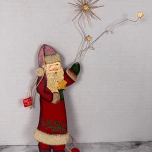 May include: A flat wooden Santa Claus ornament wearing a red coat with green leaf accents and a cream-colored fur trim. Santa holds a wire structure featuring a gold starburst and two smaller gold stars. A red bead hangs from the bottom.