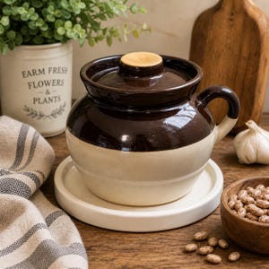 May include: A brown and cream ceramic crock with a lid and handle, on a white plate. The crock has a dark brown top and a cream base. A wooden chopping board, a garlic bulb, and a small bowl of beans are in the background.