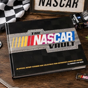 May include: A black book titled "The NASCAR Vault" with a checkered flag and a die-cast race car. The book features the NASCAR logo in red, yellow, and blue. Other items include a patch, a wrench, and a keychain with the NASCAR logo.