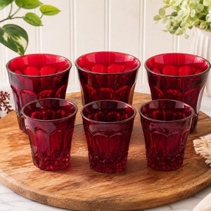 Puede incluir: Seis vasos de color rojo rubí con un diseño texturizado están dispuestos sobre una tabla de servir de madera. Los vasos tienen una forma clásica con un borde oscuro. Los vasos son de diferentes tamaños, con tres más grandes y tres más pequeños.