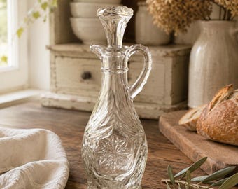 Op de afbeelding: Een heldere glazen oliekan met een decoratief bloemmotief en een handvat. De kan heeft een stop en een schenktuit. De kan staat op een houten oppervlak, met een witte doek en brood op de achtergrond.