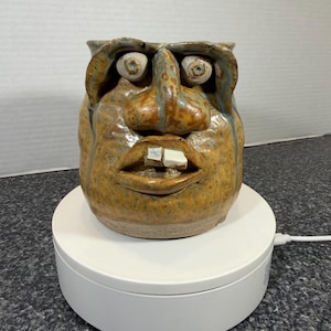 May include: A handmade ceramic mug with a face design. The mug features a tan base color with brown and blue accents, large eyes, a prominent nose, and two protruding teeth. The mug is sitting on a white rotating display.