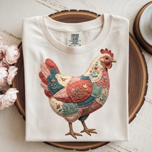 Könnte beinhalten: Cremefarbenes T-Shirt mit einem farbenfrohen Hühnerdesign im Patchwork-Stil. Das Huhn hat einen roten Kamm und Schnabel, mit Flicken in Rosa, Blau und Grün, die mit floralen Stickereien verziert sind. Das Shirt ist von Comfort Colors.