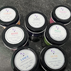 May include: Seven small, round paraffin wax candles in black tins with white labels. Each label has a different name: Coran, Shiro, Allura, Pidge, Keith, Lance, and Hunk. The candles are various colors, including orange, purple, pink, green, and beige.