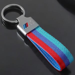 Könnte beinhalten: Ein silberner Schlüsselanhänger mit einem blau-rot-weiß gestreiften Band und einem silbernen Anhänger mit dem BMW-Logo.