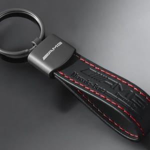 Peut inclure: Porte-clés en cuir noir avec surpiqûres rouges et le logo AMG. Le porte-clés porte la mention "Mercedes-Benz Powered By AMG".