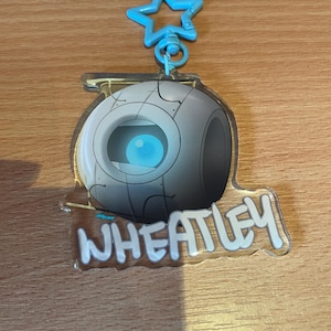 May include: A clear acrylic charm featuring a cartoon character Wheatley from the video game Portal. The charm has a blue star-shaped clasp. The character is a gray sphere with a blue eye and the name "WHEATLEY" in white.