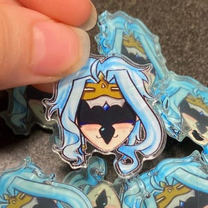 May include: A set of clear acrylic charms featuring a cartoon character with blue hair, a gold crown, and a black mask. The character has a light blue background and a light blue outline.