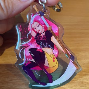 May include: A clear acrylic keychain featuring a stylized illustration of a woman with pink hair, a black top, and a yellow skirt, holding a scythe. The keychain has a silver-colored metal clasp and ring.