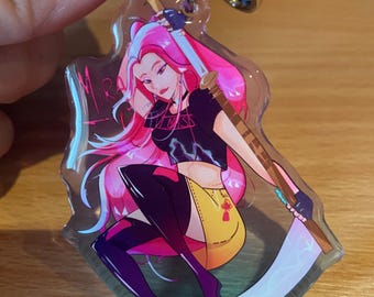 Puede incluir: Un llavero acrílico transparente con una ilustración estilizada de una mujer con cabello rosado, un top negro y una falda amarilla, sosteniendo una guadaña. El llavero tiene un cierre y un anillo de metal plateado.