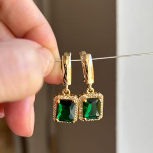 Puede incluir: Pendientes dorados con piedras preciosas verde esmeralda de corte cuadrado. Las piedras están rodeadas por un halo de piedras claras más pequeñas. Los pendientes están suspendidos por un fino alambre.