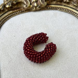 Puede incluir: Un pendiente tipo ear cuff en forma de C construido con numerosas cuentas de vidrio pequeñas de color rojo intenso. Las cuentas están densamente agrupadas para formar una banda gruesa, redondeada y texturizada que se curva en forma circular.