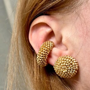 May include: Close-up of gold-colored earrings. One is a curved hoop, and the other is a round stud. Both earrings have a textured, beaded appearance, reflecting light. The earrings are worn on an ear with blonde hair in the background.