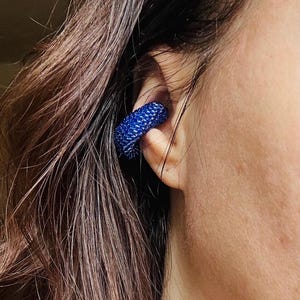 May include: A close-up of a blue beaded ear cuff. The cuff is a vibrant shade of blue and appears to be made of small, tightly packed beads. The jewelry is worn on the ear, with the wearer's skin and hair visible.