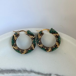 May include: A pair of beaded hoop earrings. The earrings feature a gold-colored metal clasp and are adorned with small, colorful beads in shades of green, brown, and gold. The beads are arranged in a pattern around the hoop.