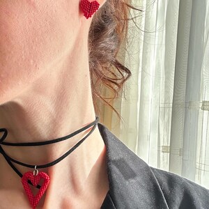May include: Red heart-shaped beaded earrings and a matching pendant necklace. The necklace features a black choker with a heart pendant. The earrings and pendant are made of small, red beads. The jewelry is worn with a black blazer.