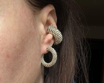 Peut inclure: Boucles d'oreilles argentées en forme de créoles avec un aspect texturé et tissé. Les boucles d'oreilles sont en forme de croissant et se portent à l'oreille. Le design présente une série de petits carrés étroitement liés, créant un motif géométrique unique.