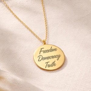 Op de afbeelding: Goudkleurige ketting met een ronde hanger. De hanger is gegraveerd met de woorden "Freedom Democracy Truth" in een sierlijk lettertype. De ketting is delicaat en dun. De achtergrond is zacht en neutraal.