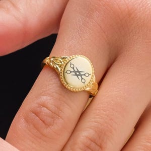 May include: A gold-colored signet ring with a circular face. The face has a cream-colored background with a black scissors design. The ring has an ornate, floral design on the band.