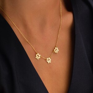 Op de afbeelding: Een goudkleurige ketting met drie pootvormige hangers. Elke hanger heeft een gegraveerde naam. De ketting wordt getoond tegen een donkere achtergrond.