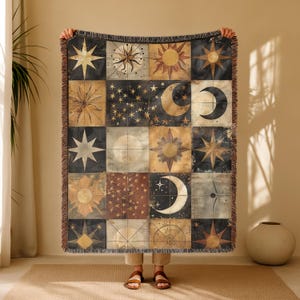May include: A woven throw blanket featuring a celestial design with suns, moons, stars, and geometric patterns in a patchwork style. The color palette includes black, gold, brown, and cream. The blanket has a fringed edge.