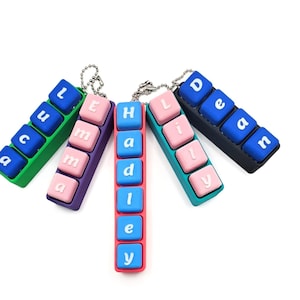May include: Five personalized keychains with names spelled out in colorful, rectangular blocks. The keychains feature names like "Luca", "Emma", "Hadley", "Lily", and "Dean", with each letter on a separate button.