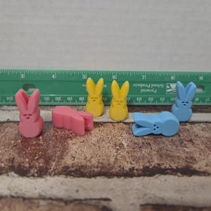 May include: A collection of colorful bunny-shaped candies in pink, yellow, and blue. The candies are arranged on a brick-patterned surface with a green ruler in the background. The candies are approximately 1 inch in height.