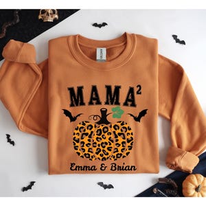 Op de afbeelding: Oranje crewneck sweatshirt met de tekst "MAMA²" in zwart boven een pompoen met een luipaardprint. De pompoen heeft een groen blad en wordt geflankeerd door twee zwarte vleermuizen. De namen "Emma & Brian" staan eronder gedrukt.