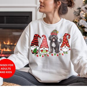May include: White sweatshirt featuring a Christmas design with three gnomes, a black dog, and the text "ME RRY CHRIS TMAS JOY". The gnomes wear red and green hats. The sweatshirt is labeled "UNISEX FOR ADULTS AND KIDS".