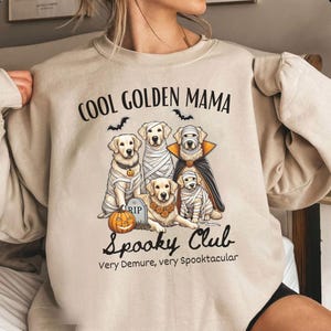May include: Beige sweatshirt with the text "COOL GOLDEN MAMA" and "Spooky Club." The graphic features five golden retrievers dressed in Halloween costumes, including a mummy, vampire, and a pumpkin. Bats are also included in the design.