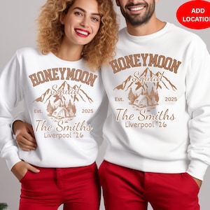 Könnte beinhalten: Zwei weiße Sweatshirts mit dem Text "HONEYMOON Squad" und "The Smiths Liverpool '26" in Braun. Das Design zeigt Berge und ein Zelt. Das Paar trägt rote Hosen.