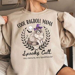 May include: Beige sweatshirt with the text "COOL RAGDOLL MAMA" above a cat wearing a witch hat, surrounded by a wreath. Below the cat, the text reads "Spooky Cult" and "Very Demure, very Spooktacular".