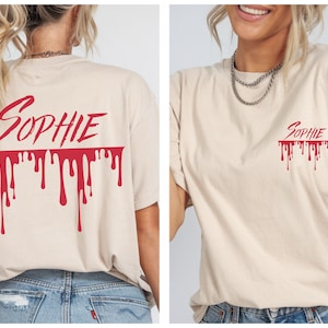 May include: A beige t-shirt featuring the name "Sophie" in red, cursive lettering above a dripping blood effect. The design is printed on the front and back. The shirt is a relaxed, unisex fit.