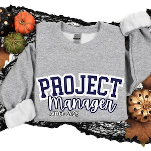 May include: Gray sweatshirt with the text "PROJECT Manager since 2025" in navy blue lettering. The text is in a varsity style font.