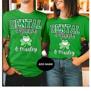 Könnte beinhalten: Zwei grüne T-Shirts mit dem Text "Dental Squad" und einem lächelnden Kleeblatt. Der Text "O'Marley" befindet sich unter dem Kleeblatt auf jedem Shirt.