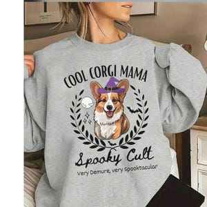 May include: A light grey sweatshirt featuring the text "COOL CORGI MAMA" above a corgi illustration wearing a purple witch hat. Below is "Spooky Cult" and "Very Demure, very Spooktacular". The design is Halloween themed.