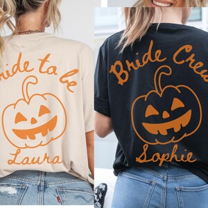 May include: Two t-shirts with Halloween designs. One is beige with "Bride to be" and a jack-o'-lantern graphic, the name "Laura" is below. The other is black with "Bride Crew", a jack-o'-lantern, and the name "Sophie".