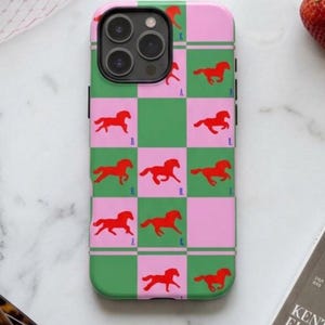 以下が含まれることがあります： ピンクとグリーンの馬柄の携帯ケース。赤い馬のシルエットが市松模様にデザインされています。大理石の表面に、ピンクの帽子、サングラス、雑誌が置かれています。