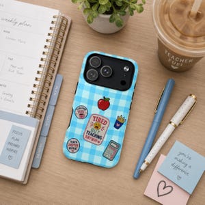May include: A light blue gingham iPhone case with various stickers, including a red apple, a coffee cup, and the text "Tired But Teaching Anyway." The case is next to a planner, pens, and a coffee cup.