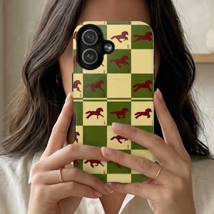 以下が含まれることがあります： 緑とベージュのチェック柄の携帯電話ケース。ケースには、栗色の馬のシルエットが繰り返されるデザインが施されています。携帯電話ケースは人の顔の前にかざされています。
