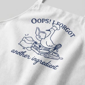 Pode incluir: Avental branco com uma ilustração azul marinho de um ganso com chapéu de chef, lendo um livro e rodeado por utensílios de cozinha. O texto diz "OOPS! I FORGOT another ingredient."