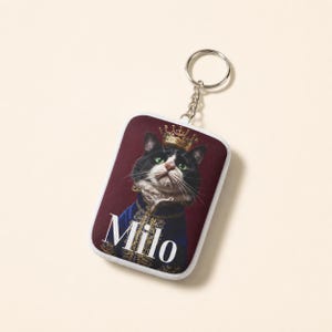 May include: A rectangular keychain featuring a portrait of a black and white cat wearing a gold crown and a blue ornate jacket. The cat is set against a dark red background with the name Milo printed in white capital letters at the bottom.