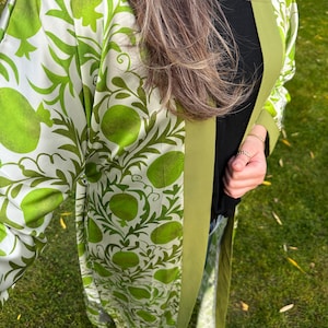 May include: A long, flowing kimono-style robe with a white background and a vibrant green pomegranate pattern. The robe has a solid green trim and is made of a lightweight fabric. The garment is worn over a black top and jeans.