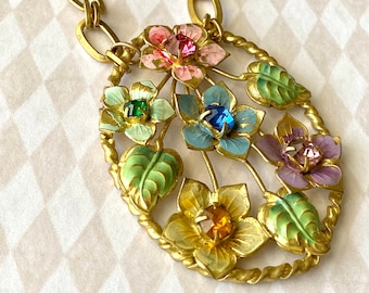 May include: A gold chain necklace with a pendant featuring an oval-shaped design with five enamel flowers and green leaves. The flowers are decorated with colorful rhinestones.