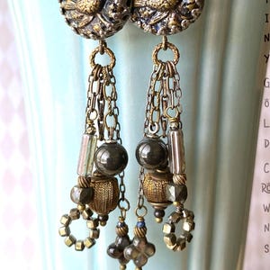 May include: Antique-style dangle earrings with a bird design. The earrings feature a gold-colored bird on a round, textured base. Below the bird are chains, beads, and decorative elements. The earrings are suspended from a hook. The text "TINY GOLD CROWS" is visible.