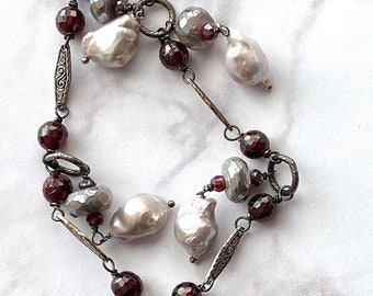 May include: A long silver chain necklace with a silver clasp. The necklace is adorned with multiple baroque pearls and dark red faceted beads.