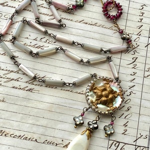 May include: A white and gray shell necklace with a large, ornate, gold-toned pendant. The pendant features a cluster of raised, gold-toned flowers and is surrounded by a white shell. The necklace is accented with small, pink and gray beads.