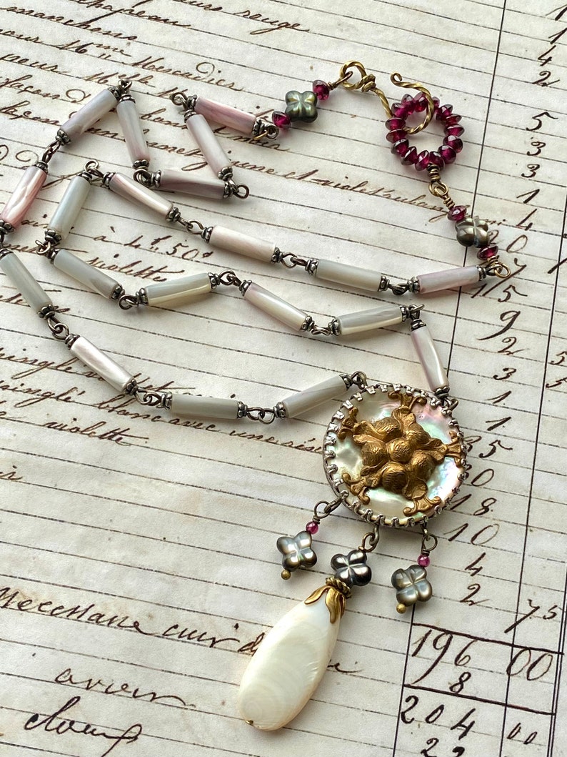 May include: A white and gray shell necklace with a large, ornate, gold-toned pendant. The pendant features a cluster of raised, gold-toned flowers and is surrounded by a white shell. The necklace is accented with small, pink and gray beads.