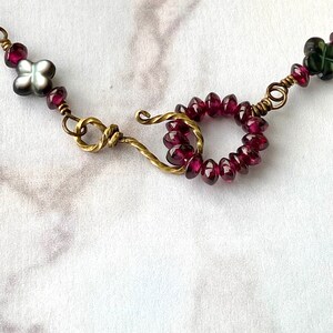 May include: A close-up of a necklace with a brass clasp and a circle of dark red beads. The necklace also features a small white flower bead.